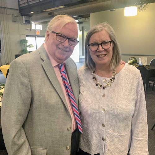 The night's Master of Ceremonies, Paul Scott, stands smiling in a grey suit with a pink dress shirt. He wears square frame glasses. Net to him in the photo is WRFN Executive Director, Sue Simpson, she wears a white buttoned down blouse that is textured. She is smiling and wears square framed glasses and a necklace with pedants